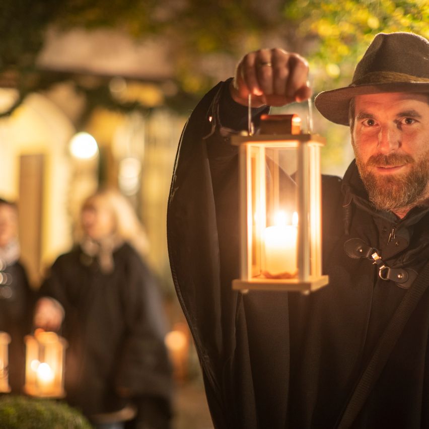 Ein Mann in Kostüm hält draußen bei Nacht eine brennende Laterne, mit anderen im Hintergrund.