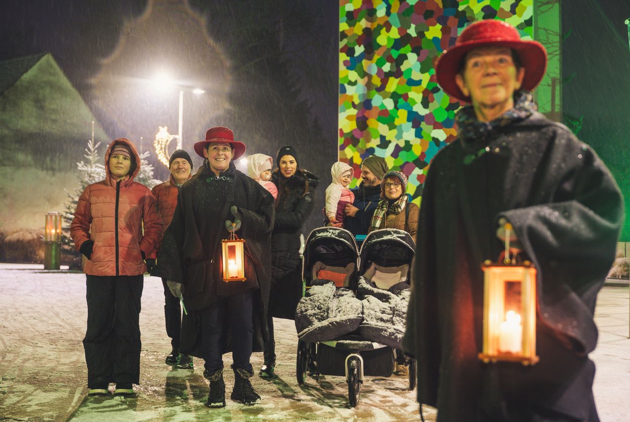 Eine Gruppe von Menschen in Mänteln und Mützen, die Laternen halten, steht im Schnee. Einige tragen Babys in Kinderwagen. Eine bunte Wand befindet sich hinter ihnen.