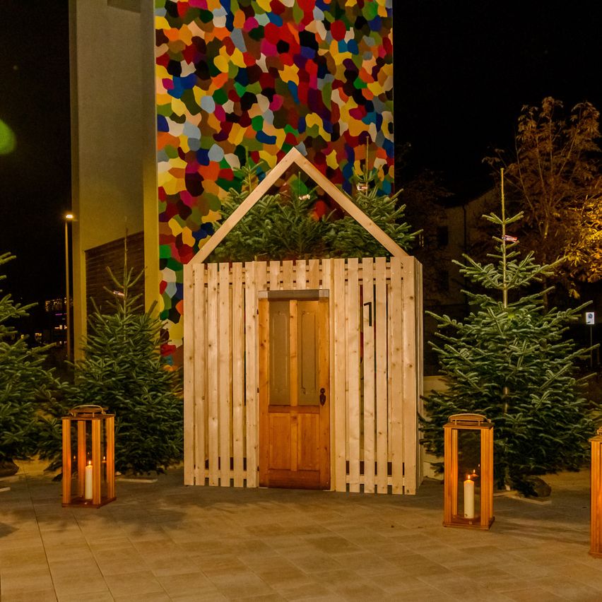 Ein kleines Holzhaus steht bei Nacht, geschmückt mit Weihnachtsbäumen und Kerzen, vor einer farbenfrohen, lebhaften Wand.
