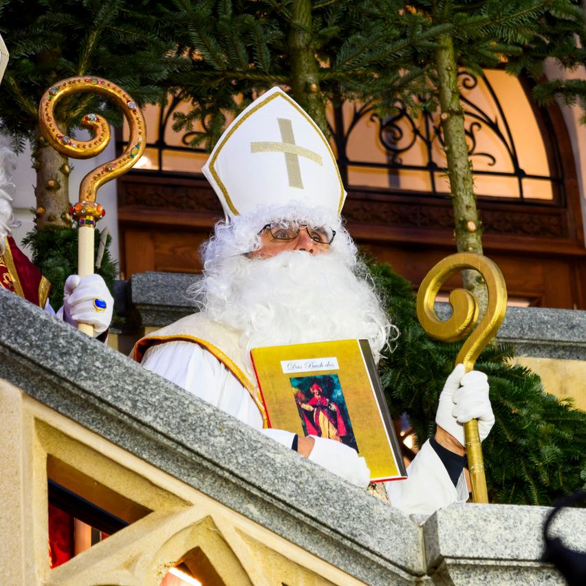Eine als Bischof verkleidete Person hält ein Buch und einen Stab auf einem Balkon, mit Tannenbäumen und einem Gebäude im Hintergrund.