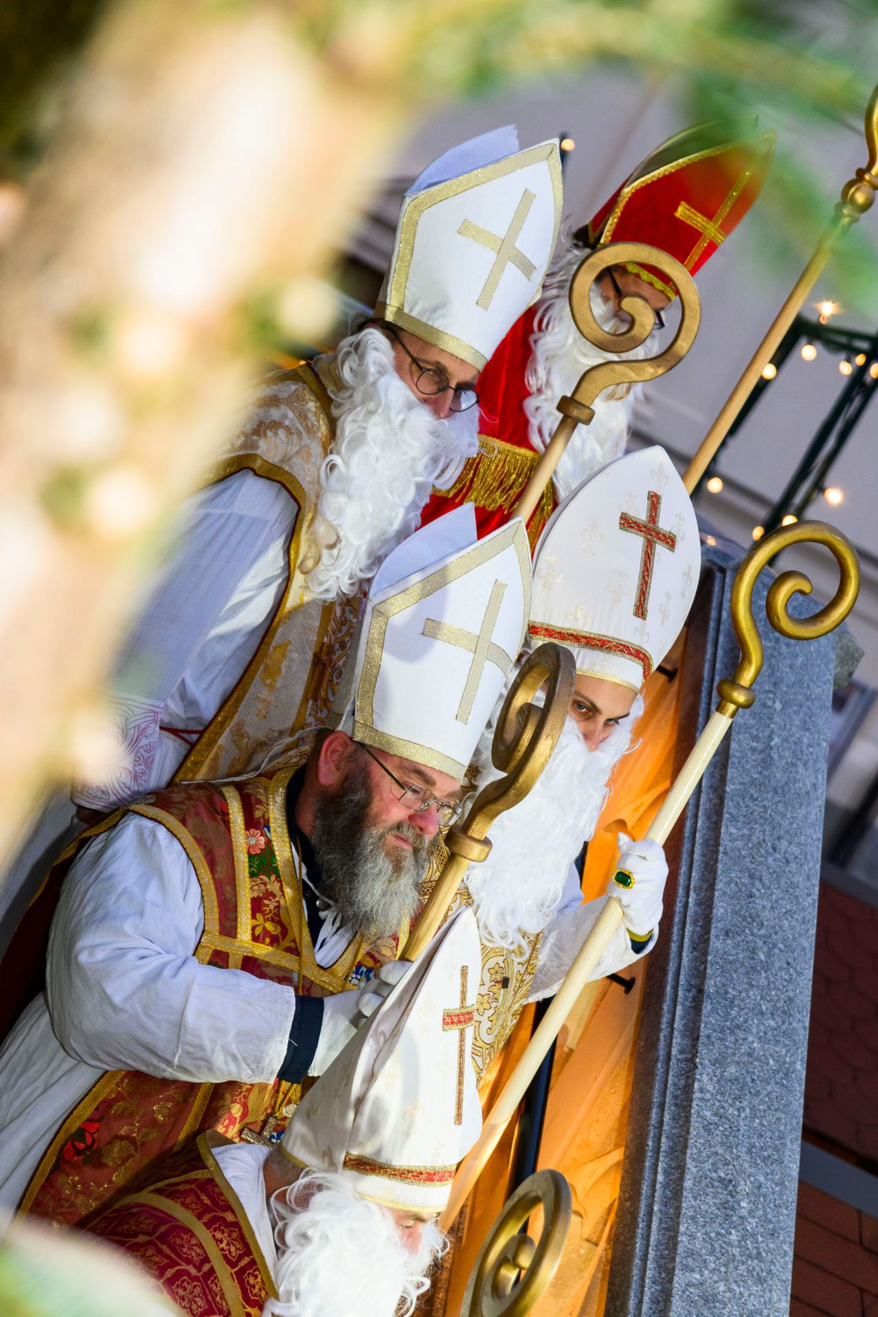 Drei Männer in weißen Gewändern und Mitras, die Bischöfen ähneln, stehen eng beieinander, einer hält einen goldenen Stab. Sie sind in einer Kirche, mit dekorativen Lichtern an der Decke.