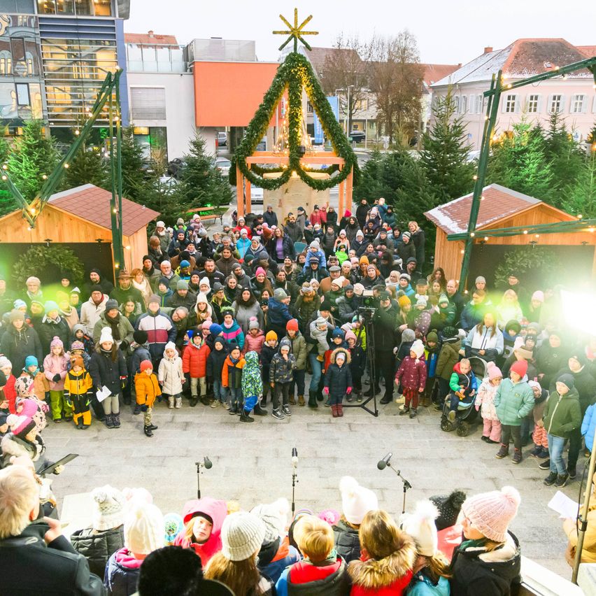 Eine Menschenmenge, darunter Kinder, versammelt sich in einer festlichen Außenumgebung mit Weihnachtsbäumen und Dekorationen. Ein großer beleuchteter Weihnachtsbaum steht in der Mitte. Einige tragen Mützen und Winterkleidung.