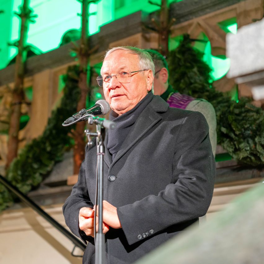 Ein älterer Mann mit Brille und schwarzem Mantel steht an einem Mikrofon und hält vermutlich eine Rede. Hinter ihm befinden sich grüne Lichter und Weihnachtsdekorationen.