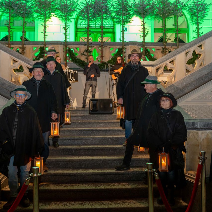 Eine Gruppe von Menschen in schwarzen Roben und Hüten geht eine Treppe hinauf und hält brennende Laternen. Die Treppe ist mit grünen Lichtern dekoriert. Ein Mann steht mit einem Mikrofon an der Seite.
