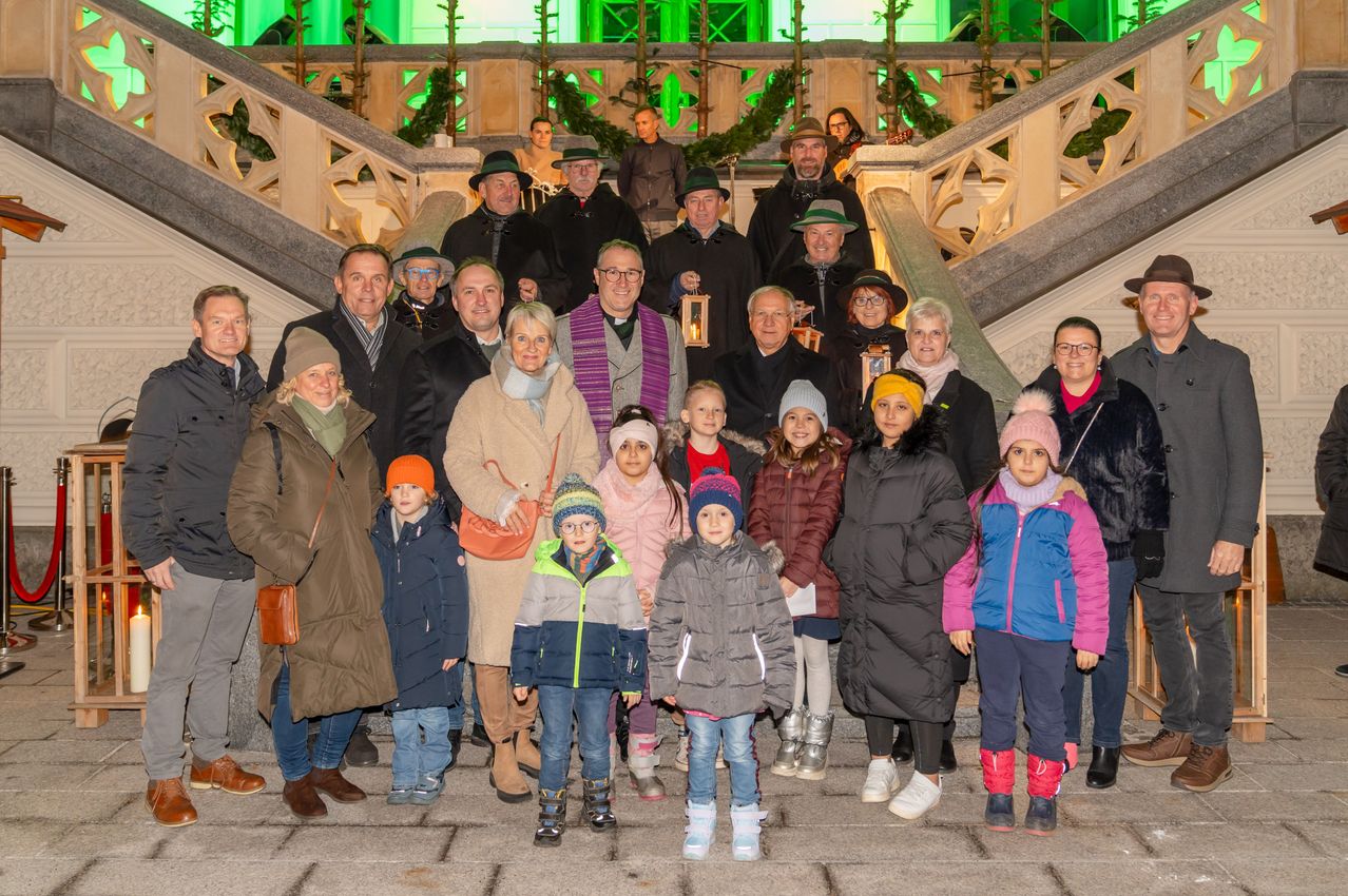 Eine Gruppe von Menschen steht auf einer Treppe, einige mit Winterkleidung und Mützen. Im Hintergrund befindet sich eine steinerne Struktur mit dekorativen grünen Lichtern.