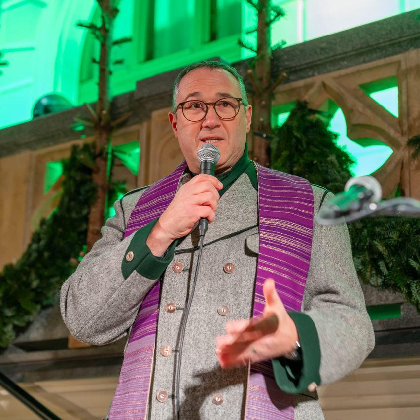 Ein Mann steht vor einem Mikrofon mit einer violetten Stola, hält es mit beiden Händen und macht Gesten, wahrscheinlich während einer religiösen Veranstaltung. Dahinter befindet sich eine grün beleuchtete architektonische Gestaltung mit Pflanzen.