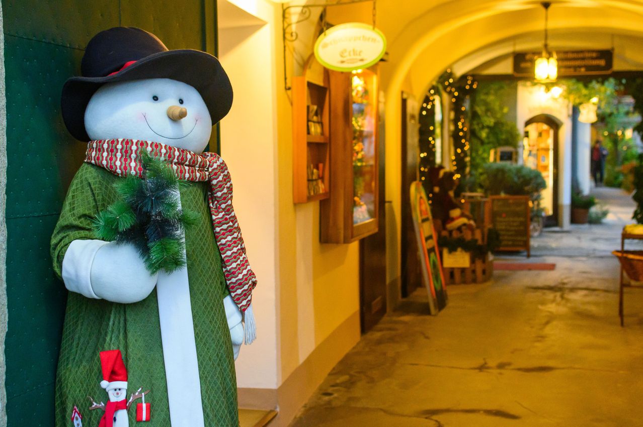 Ein Schneemann steht lächelnd draußen, mit einem grünen Kleid, Schal und einem Weihnachtsbaum. Dahinter steht ein Schild mit der Aufschrift 'Ecke'. Ein Weg führt zu einem gewölbten Eingang.