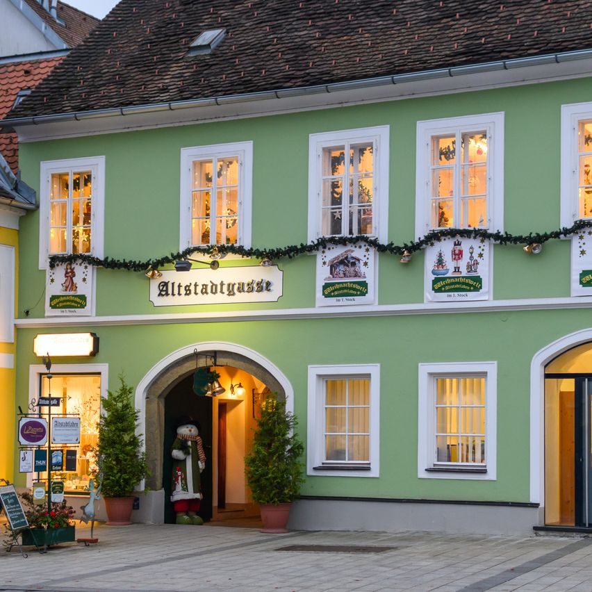 Die Vorderseite eines grünen Gebäudes mit einem Schild, das Altstadtgasse liest. Es hat viele Fenster, einen beleuchteten Bogeneingang und eine Weihnachtsdekoration darüber. Topfpflanzen und ein Schild stehen auf dem Bürgersteig.