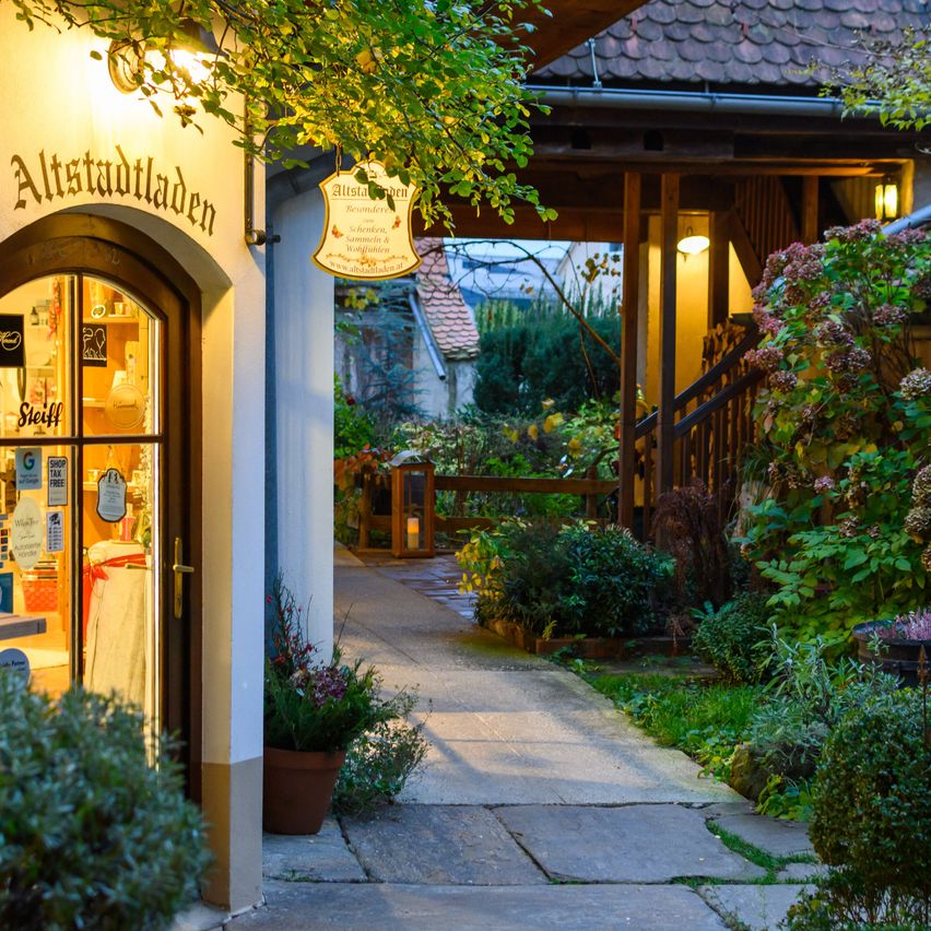 Ein Weg führt zu einem Gebäude mit 'Altstadtladen' Schild. Das Gebäude hat eine Glastür mit Gegenständen im Inneren und einem Topfpflanzen. Der Weg wird von Pflanzen und einer Lampe flankiert.