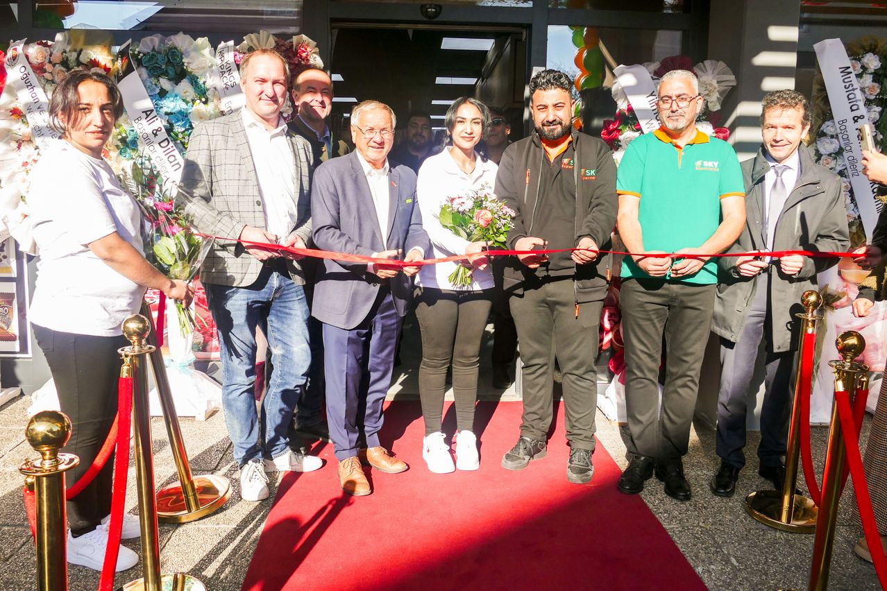 A group of people stands on a red carpet, cutting a ribbon. A woman holds flowers, and men wear business suits. Behind them, two men in polo shirts are labeled SKY. The scene is outdoors.