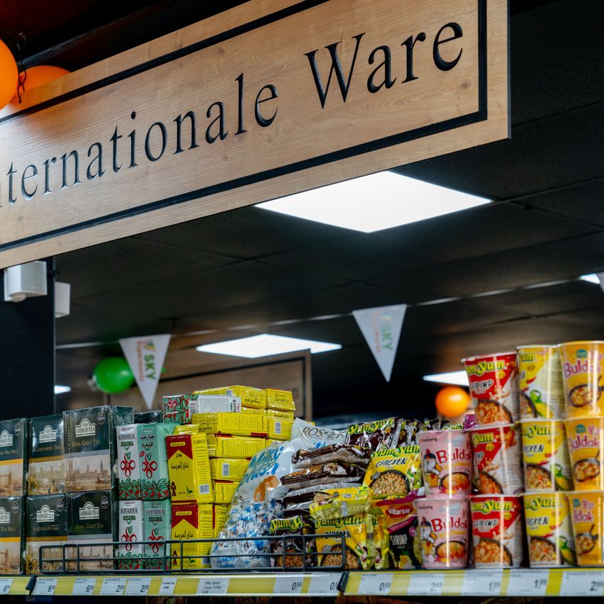 A store with a wooden sign that reads 'Internationale Ware' has various products on display, including tea boxes, snacks, and packaged noodles, under ceiling lights.