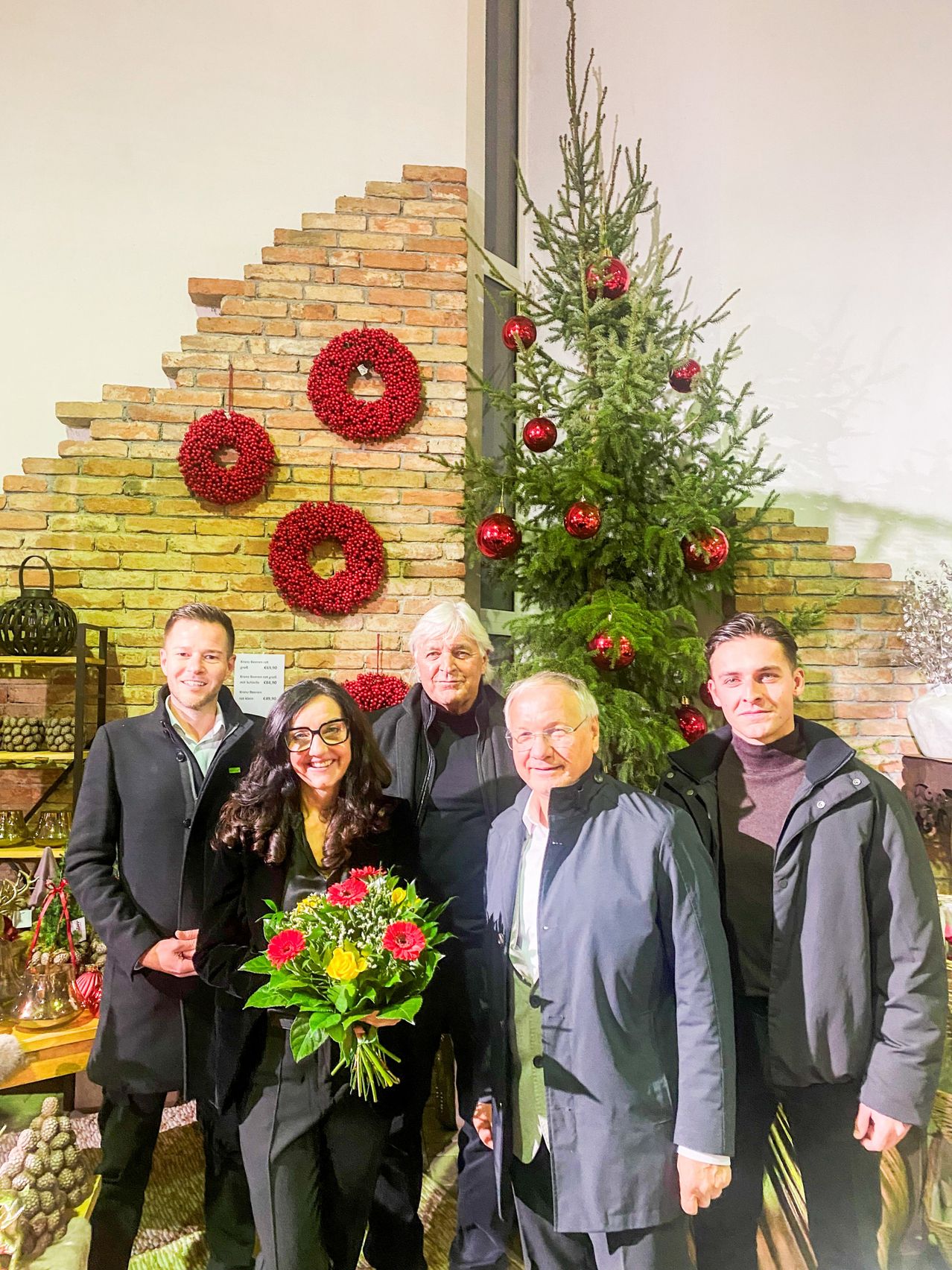 Five individuals stand in front of a decorated Christmas tree with ornaments, smiling for a photo. A woman in the center holds a bouquet of flowers. Behind them, a brick wall has hanging wreaths, and a window reflects the tree.