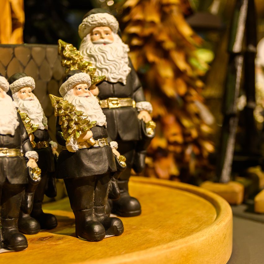 Four black Santa Claus figurines with white beards and gold accessories stand on a wooden plate. Behind them is a Christmas tree and a blurry background.