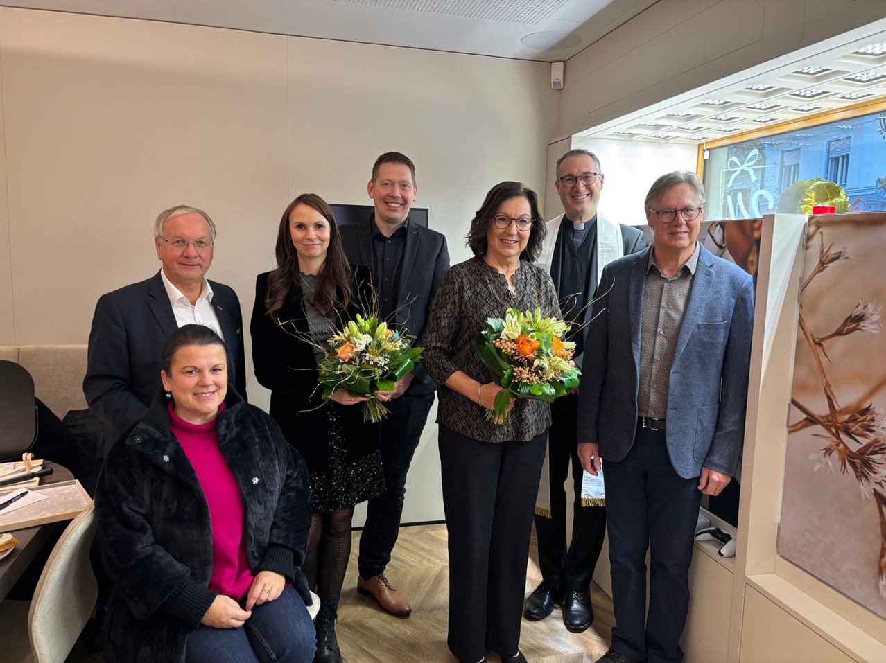 Eine Gruppe von sechs Erwachsenen, darunter zwei Frauen, steht und sitzt zusammen in einem Raum, lächelt und hält Blumensträuße. Sie sind feierlich gekleidet. Hinter ihnen sind eine Wand und ein Glasfenster zu sehen.