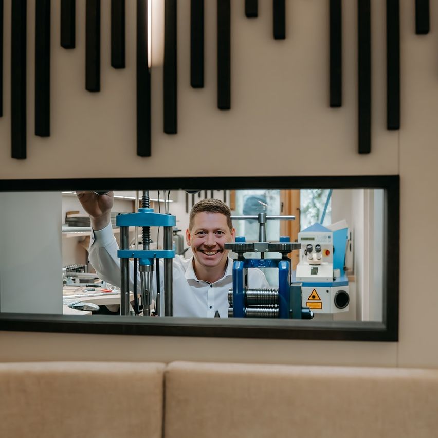 Ein Mann in einem weißen Hemd lächelt vor einer Maschine in einem Labor. Ein Spiegel reflektiert die Szene.