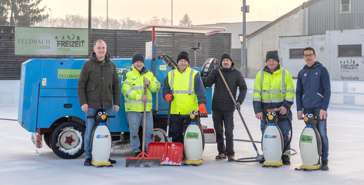 Fünf Männer, in Winterkleidung gekleidet, stehen auf einer verschneiten Eisbahn mit Reinigungsgeräten. Zwei Pinguine, einer mit Besen und Schaufel, stehen auf jeder Seite. Dahinter ist ein blauer Schneepflug geparkt.