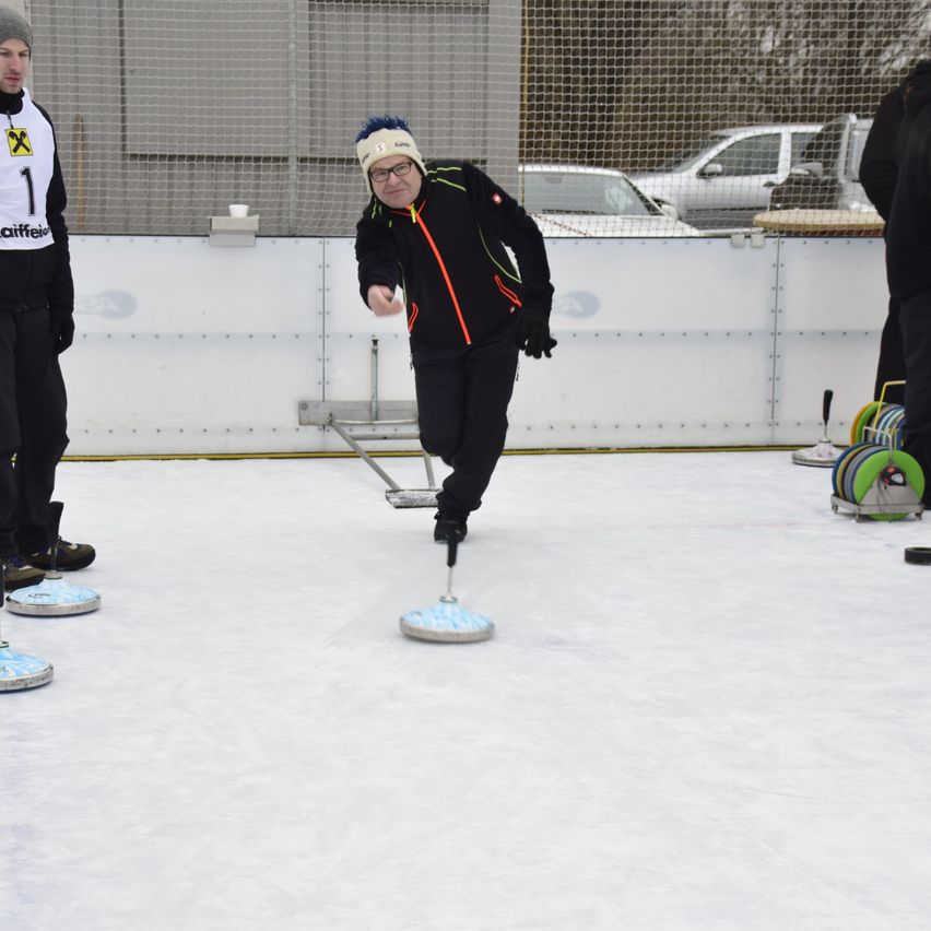Ein Mann auf einer Eisbahn wirft einen Puck, während zwei andere in der Nähe stehen. Er trägt eine Winterjacke und eine Mütze. Auf dem Eis befinden sich vier blaue Ziele.