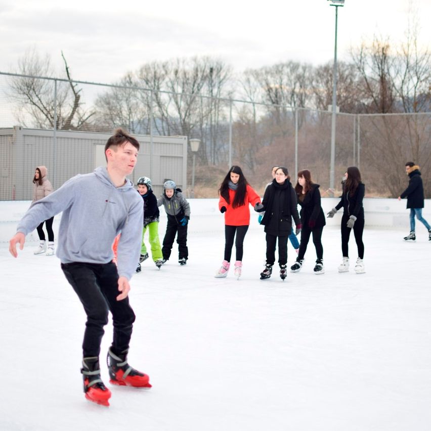 Eine Gruppe von Menschen skat auf einer Freiluft-Eisbahn. Ein Mann in Grau fährt vorwärts, während andere hinter ihm her folgen. Der Himmel ist bewölkt.