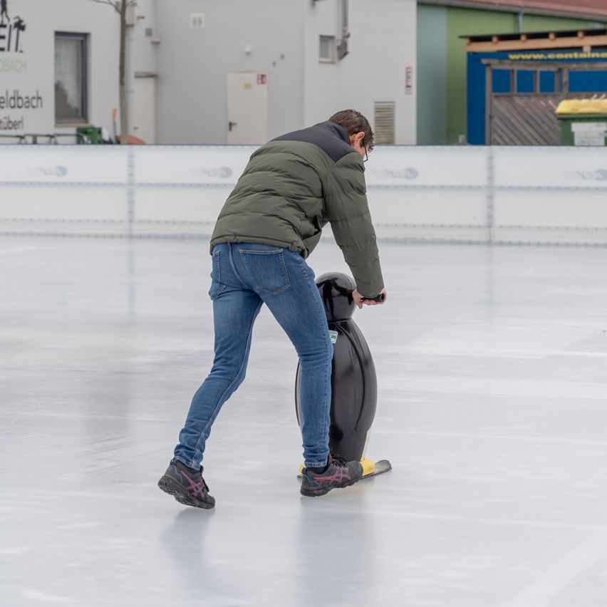 Ein Mann eisskat auf einer Eisbahn im Freien und hält einen pinguinförmigen Eisschuh in der rechten Hand.