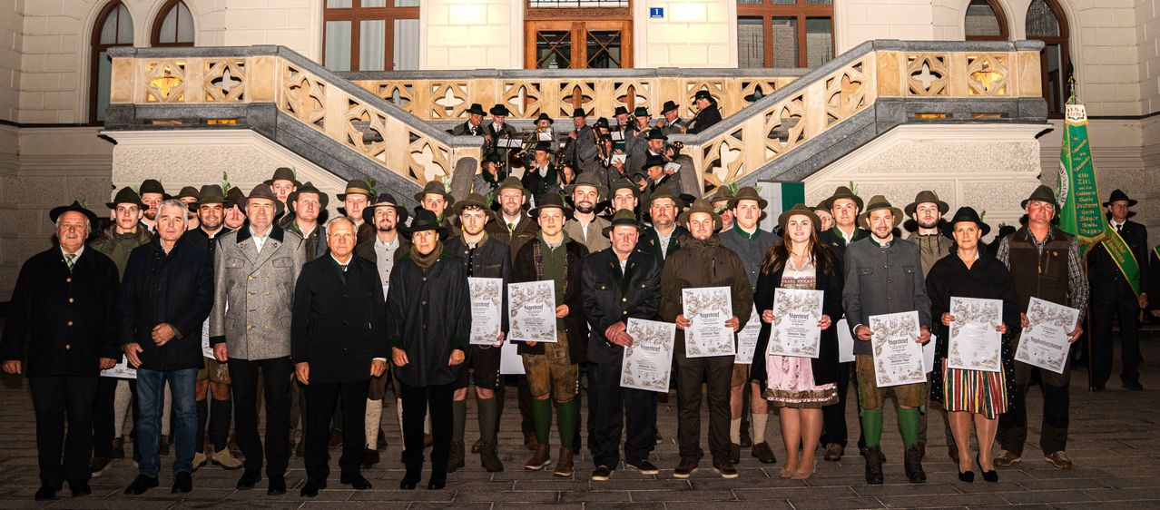 Eine Gruppe von Menschen in traditioneller Kleidung, einschließlich Hüten und Stiefeln, steht auf einer Treppe und hält Urkunden. Dahinter spielt eine Band Instrumente.