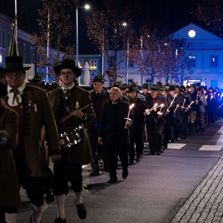 Eine nächtliche Prozession von Menschen in traditioneller Kleidung und Hüten, die Kerzen halten, zieht unter Straßenlaternen die Straße hinunter.
