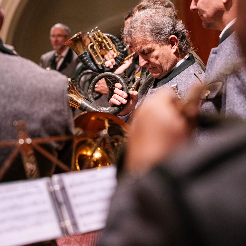 Ein älterer Mann in Uniform, der eine Trompete hält, scheint in einem ernsten Moment mit anderen um ihn herum zu sein. Musikinstrumente und Papiere sind im unscharfen Hintergrund vorhanden.