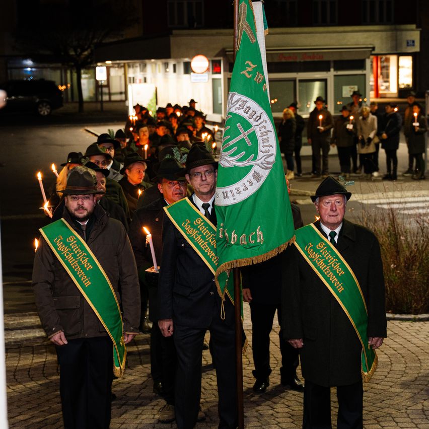 Eine Gruppe von Menschen hält Kerzen und trägt grüne Schärpen, möglicherweise bei einer Gedenkveranstaltung. Ein Mann in Anzug hält eine grüne Flagge vor sich. Im Hintergrund sind weitere Menschen mit Kerzen zu sehen.