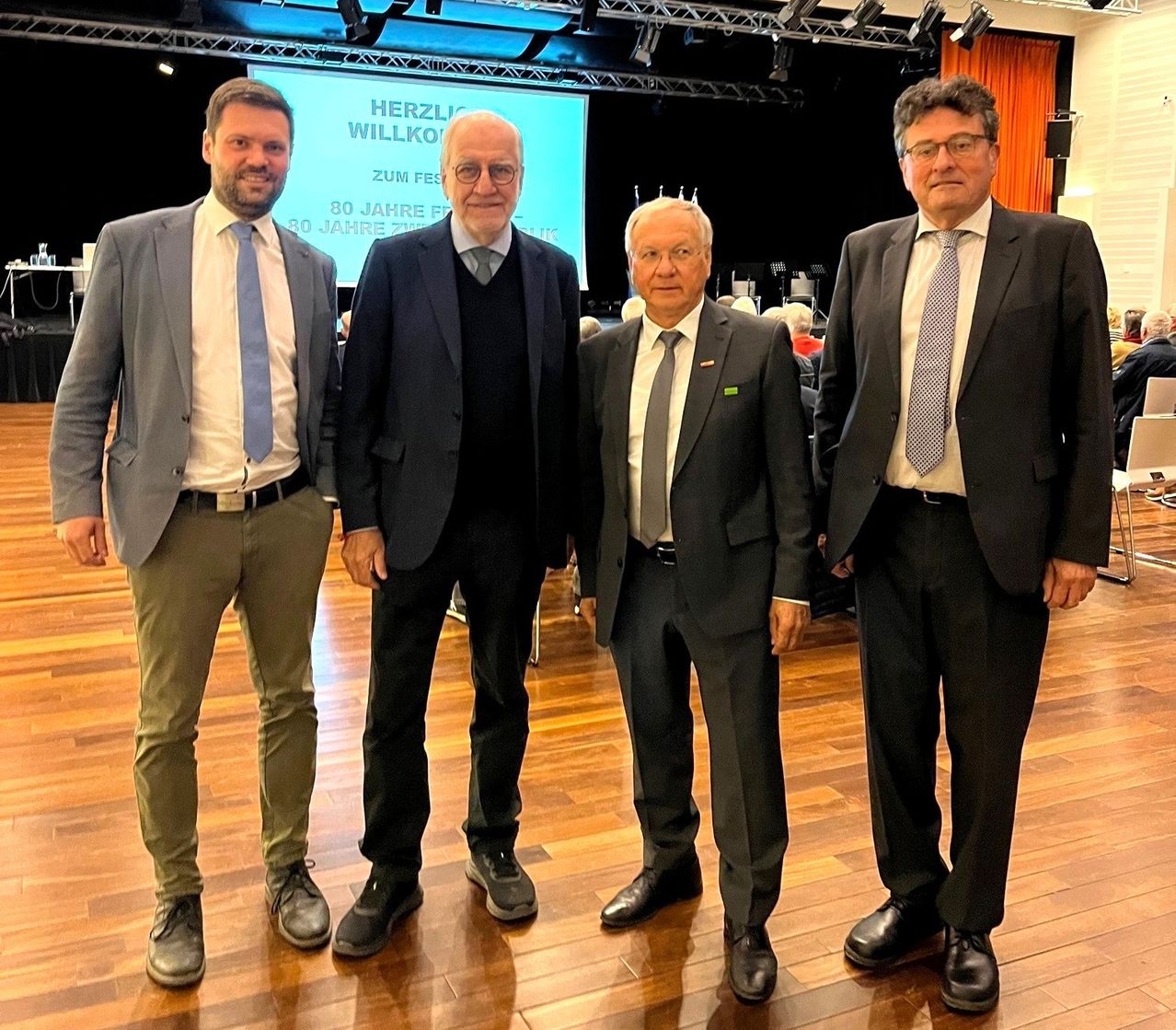 Vier Männer in Anzügen und Krawatten stehen zusammen auf einem Holzboden. Hinter ihnen zeigt eine Leinwand eine Präsentation an.