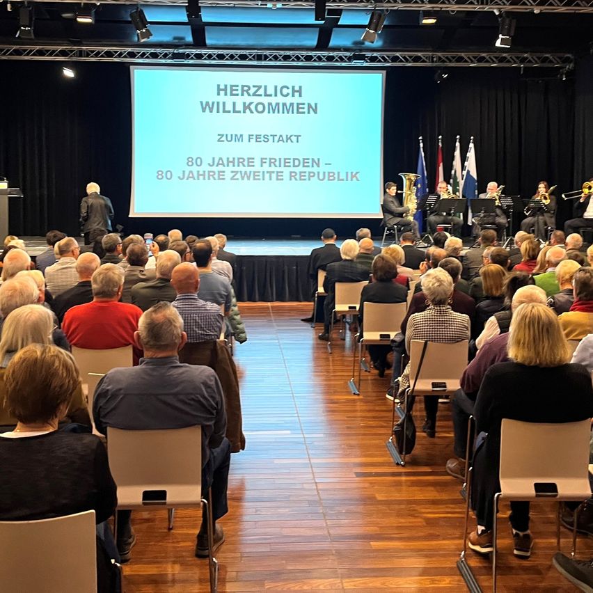 Ein Publikum sitzt in Stühlen bei einem Konzert. Auf der Bühne wird eine Leinwand mit deutschem Text angezeigt, der die Besucher zu einer Feier von 80 Jahren Frieden und der zweiten Republik begrüßt.