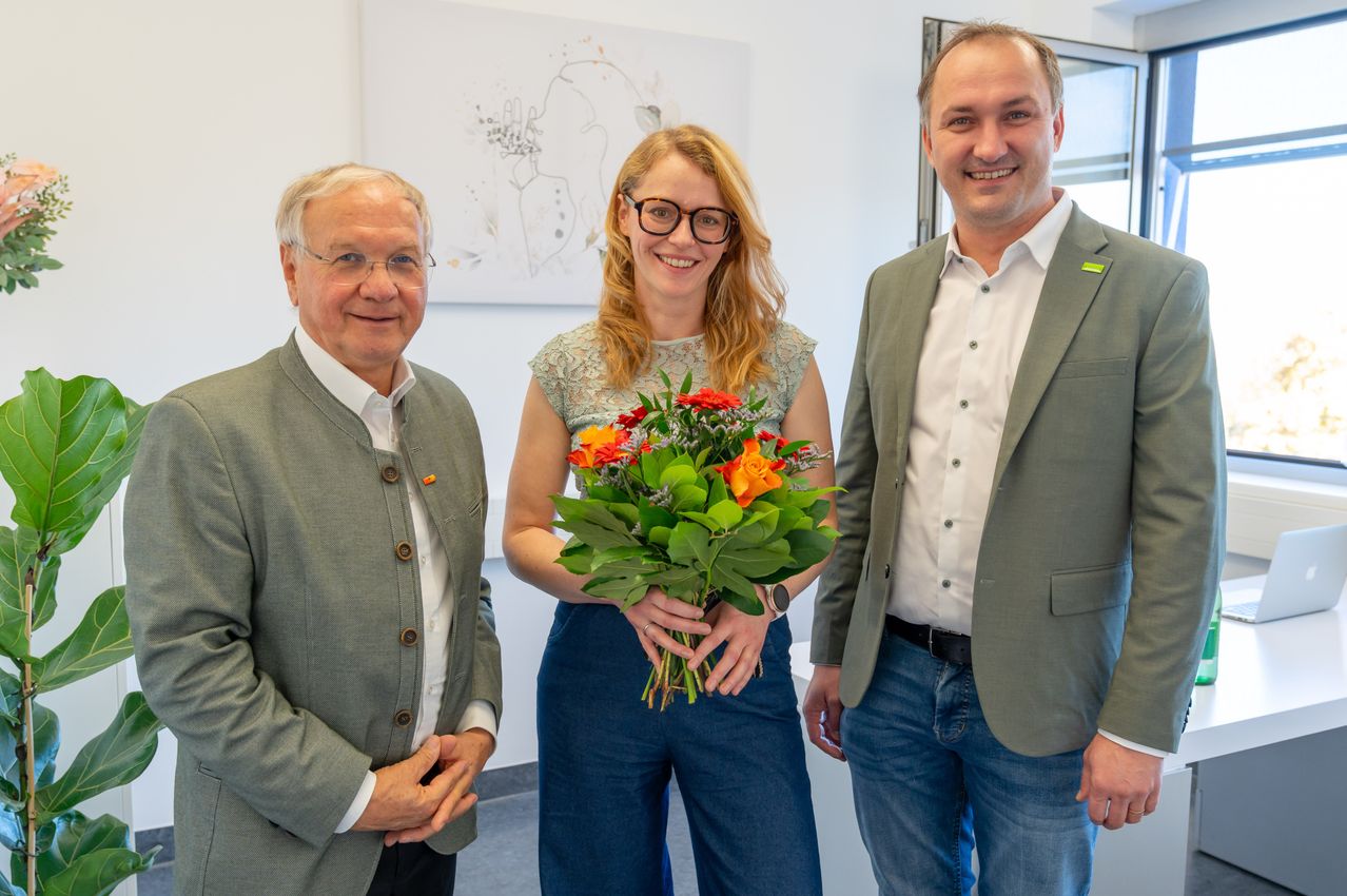 Drei Personen stehen lächelnd nebeneinander. Eine Frau hält einen Blumenstrauß. Ein Mann links trägt einen grauen Blazer. Hinter ihnen ist eine weiße Wand mit einem Gemälde.