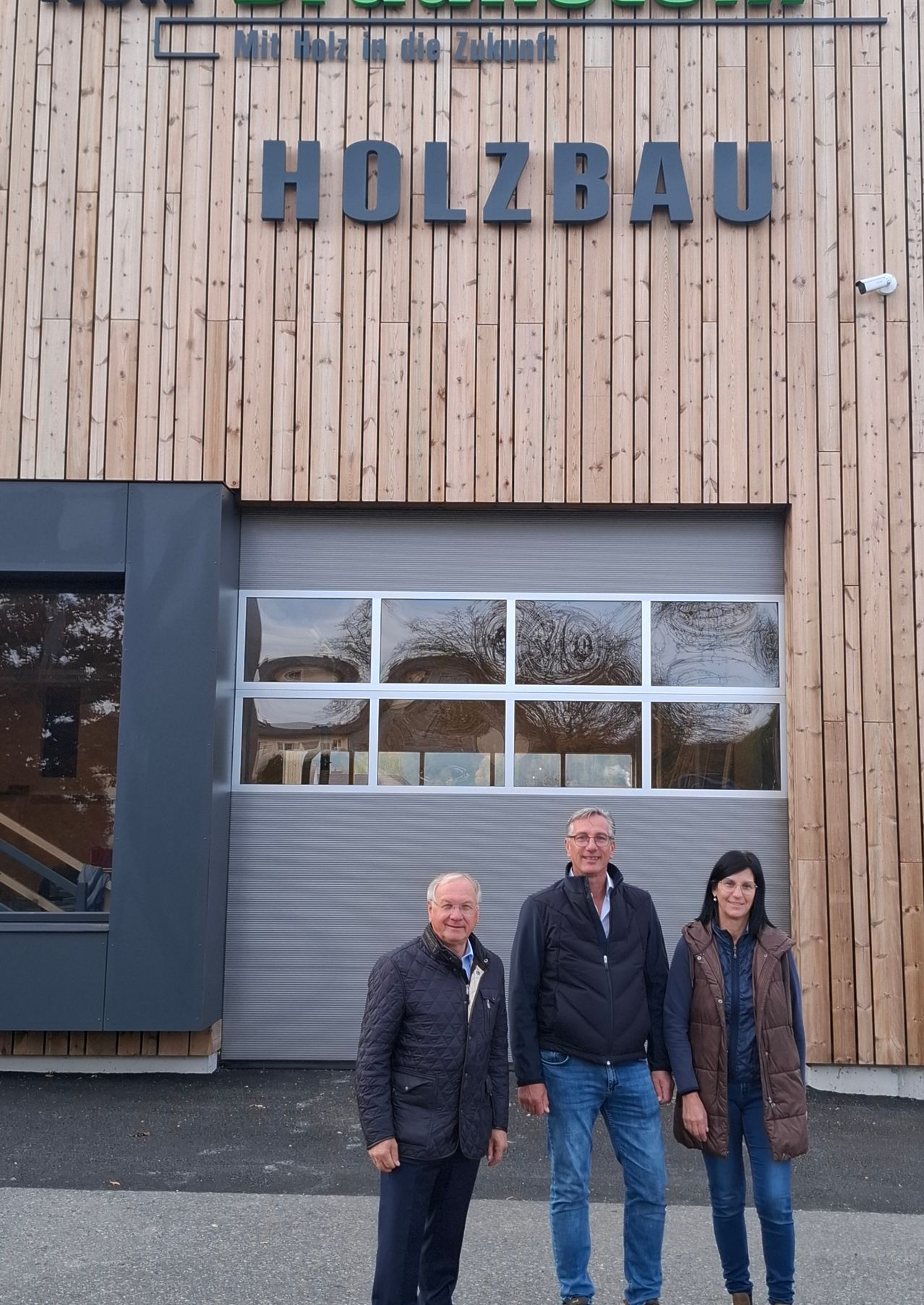 Drei Personen stehen vor einem Gebäude mit einem Schild, das 'Holzbau' steht. Sie lächeln für ein Foto. Das Gebäude hat Holzwände und eine graue Garagentür.