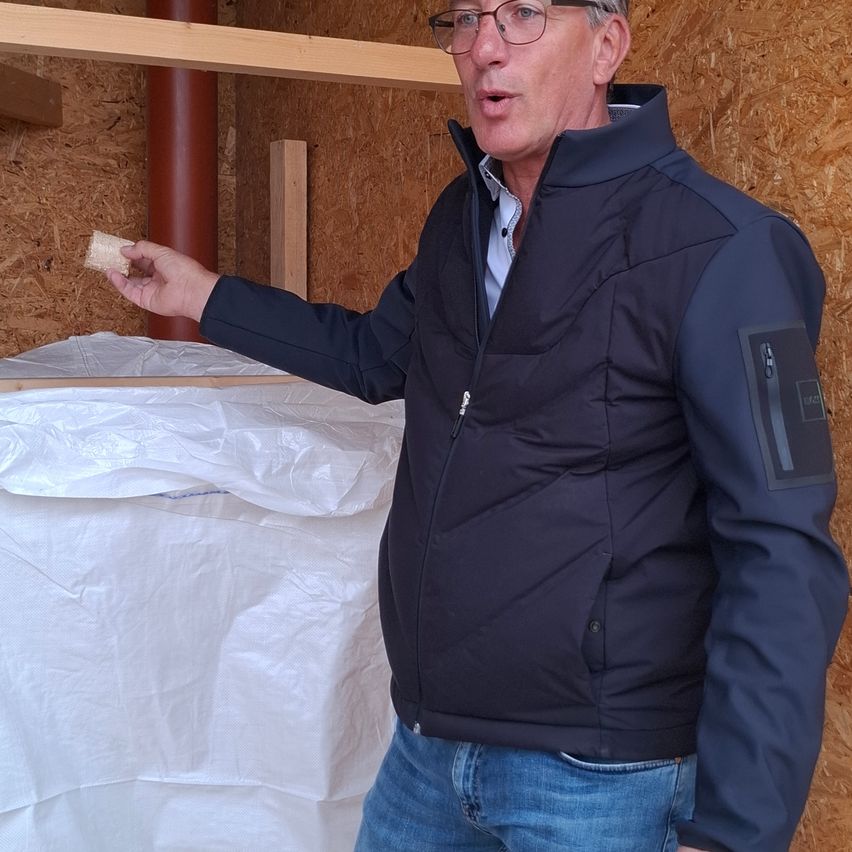 Ein Mann mit Brille und einer dunklen Jacke spricht in einem Raum mit Holzwänden. Er hält ein kleines Stück Brot und gestikuliert mit der anderen Hand.