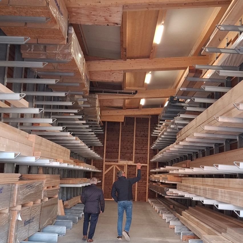 Zwei Männer gehen einen Gang in einem Lagerhaus mit Holzregalen und Balken entlang. Sie tragen beide Jacken und Sneakers.