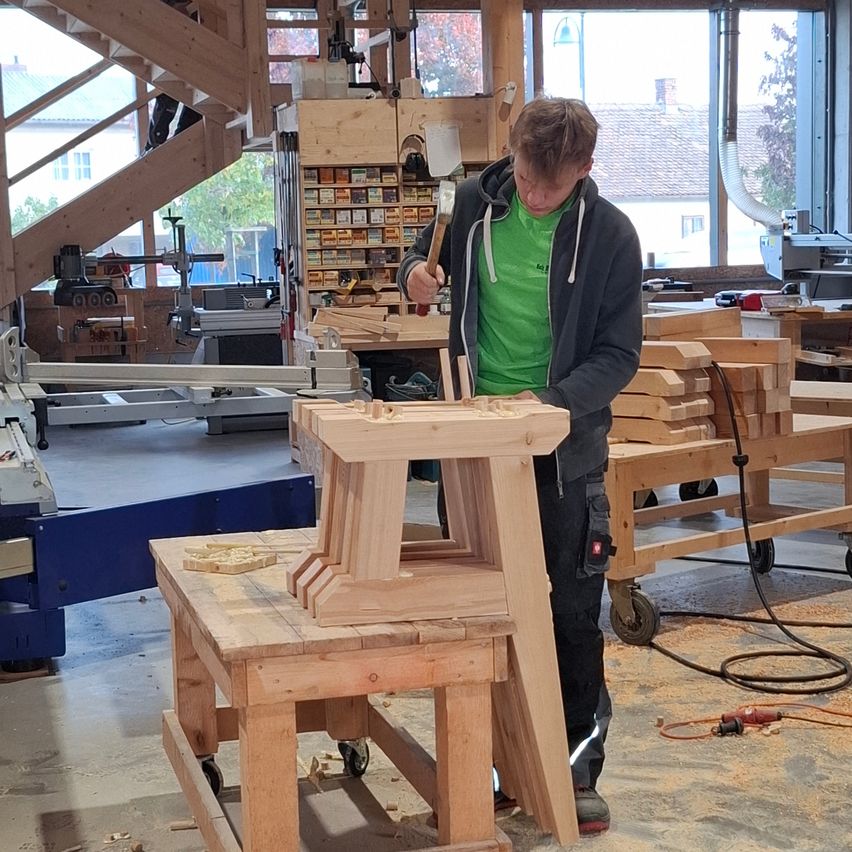 Ein junger Mann in einer Werkstatt baut einen hölzernen Stuhl zusammen. Er trägt ein grünes Shirt und schwarze Hose. Verschiedene Holzteile und Werkzeuge sind um ihn herum.