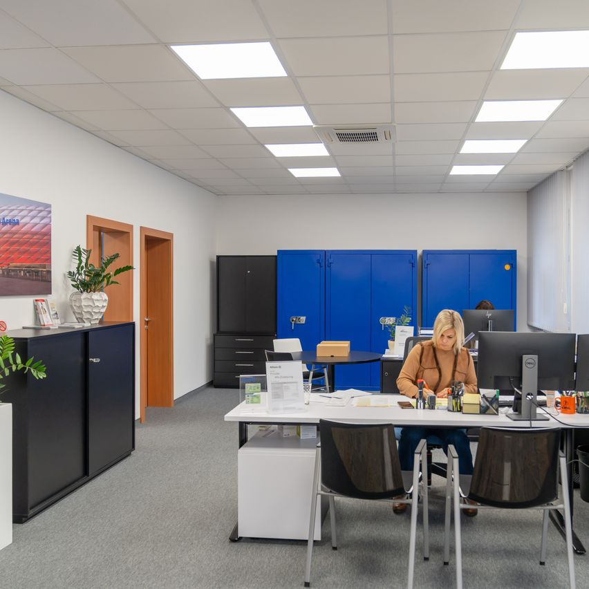 Ein Büro mit einer Frau, die an einem Schreibtisch mit Computer und verschiedenen Gegenständen sitzt. Blaue Schränke und Holztüren im Hintergrund. Der Raum hat grauen Teppich und Deckenleuchten.