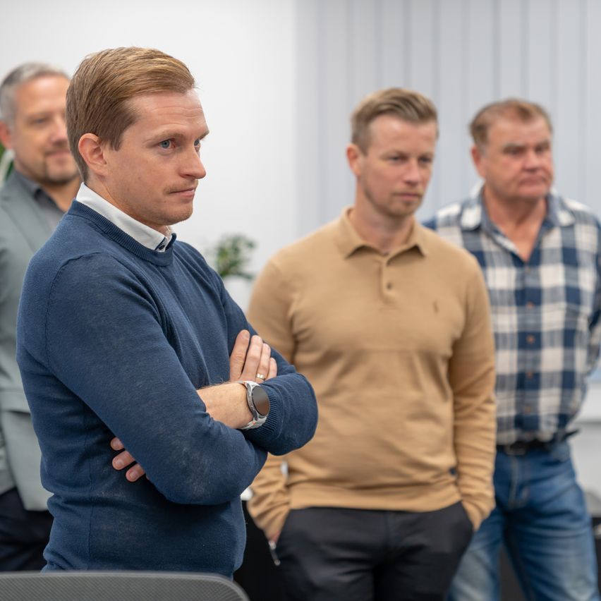 Vier Männer in einem Raum, einer mit verschränkten Armen. Hinter ihm ein Mann in einem braunen Hemd. Ein weiterer Mann im karierten Hemd auf der rechten Seite. Dahinter eine weiße Wand.