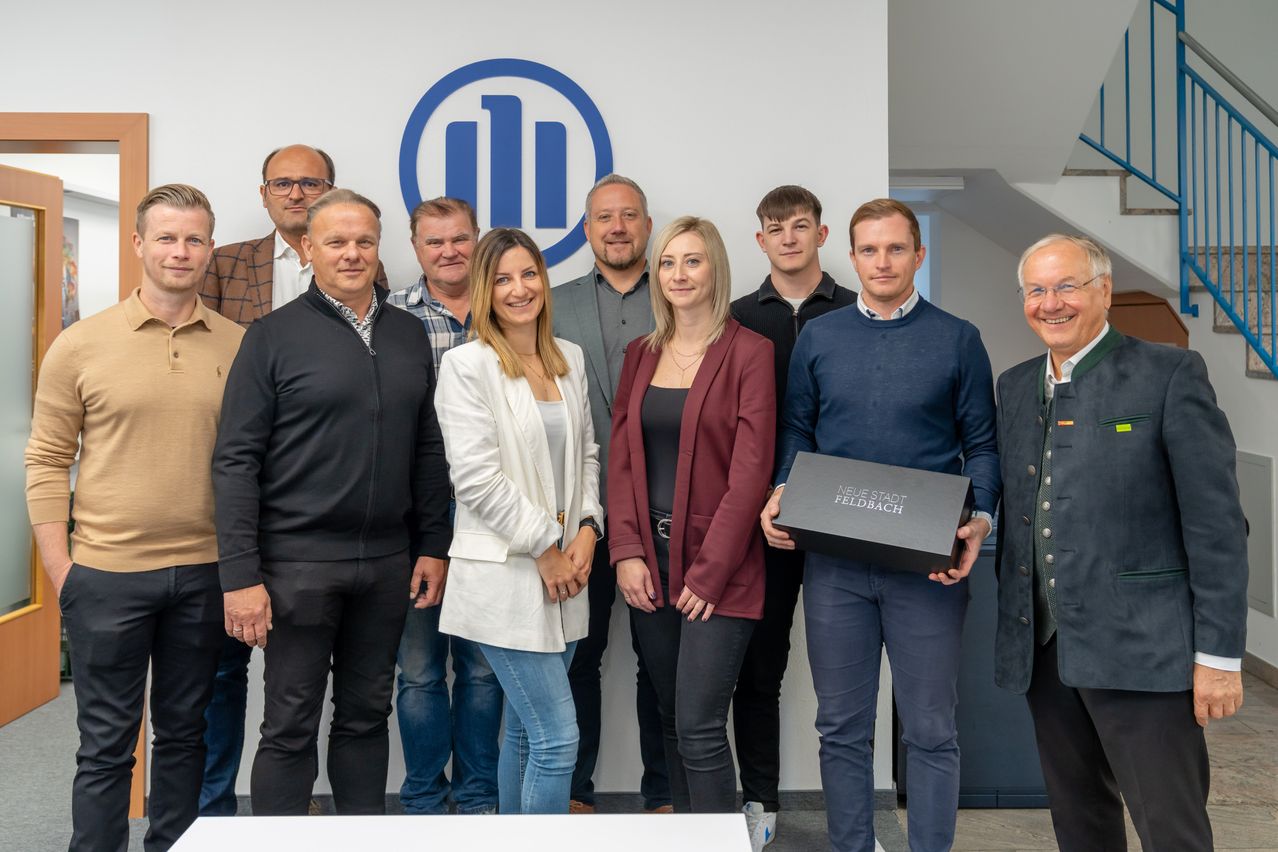Eine Gruppe von Menschen posiert für ein Foto in einem Büro mit einem blauen Logo im Hintergrund. Ein Mann hält eine schwarze Box mit den Worten 'Neue Stadt Feedback'.