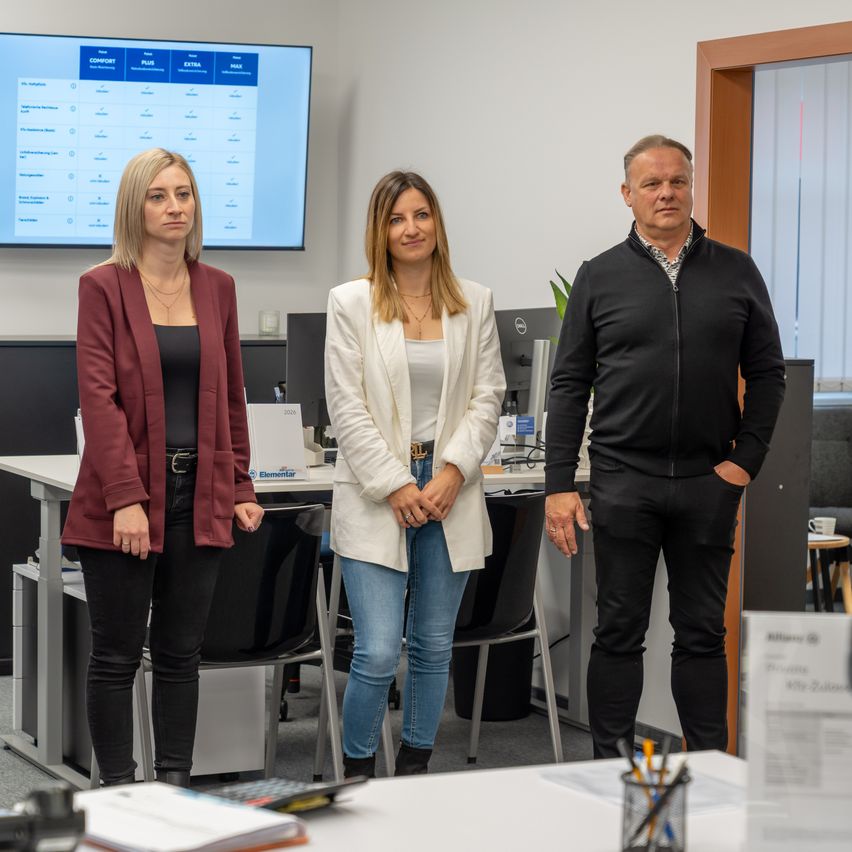 Drei Personen stehen in einem Büro, eine Frau mit blonden Haaren, eine weitere mit langen blonden Haaren und ein Mann. Sie stehen vor einem Schreibtisch, im Hintergrund sind Monitore und Pflanzen zu sehen.