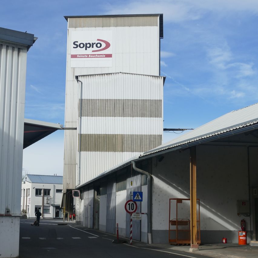 Ein hohes Sopro-Gebäude mit Logo an der Seite befindet sich in einem Fabrikgelände. Es gibt ein Geschwindigkeitslimit-Schild und eine Person, die geht.