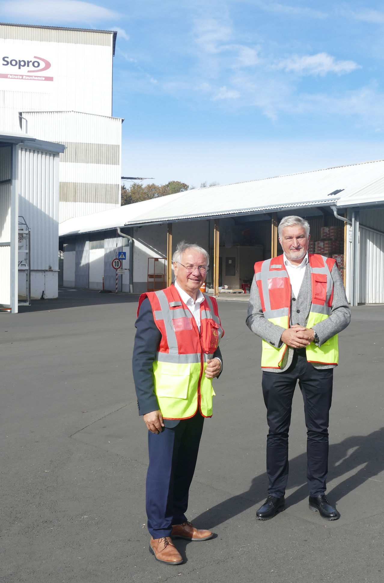 Zwei Männer in Sicherheitswesten stehen vor einem Lagerhaus, einer mit einem Werkzeug in der Hand und der andere lächelnd. Dahinter befindet sich ein Gebäude mit einem Geschwindigkeitsschild.
