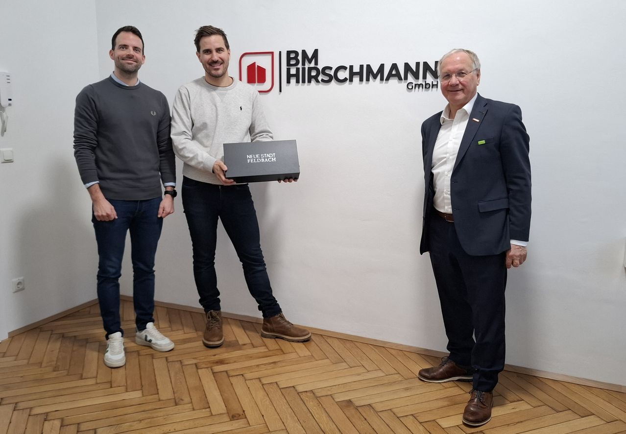 Three men stand in front of a wall with a BM Hirschmann GmbH logo. One holds a box, smiling with two others.