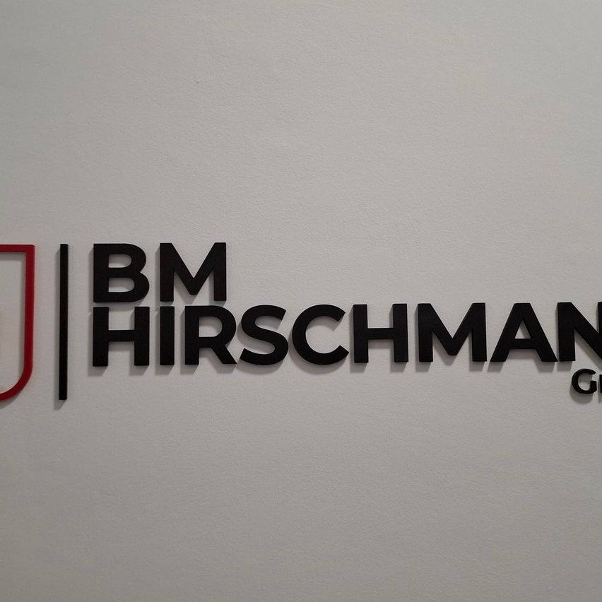 A white wall with a logo for BM Hirschman Group, featuring a red and black logo with 'BM' and 'HIRSCHMAN' in bold black font.