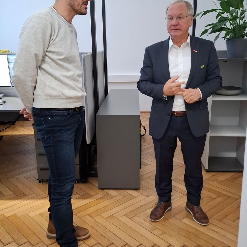 Two men in a room, one in a suit, the other in casual wear, stand facing each other. Behind them is a desk with a computer monitor, a plant, and shelves.