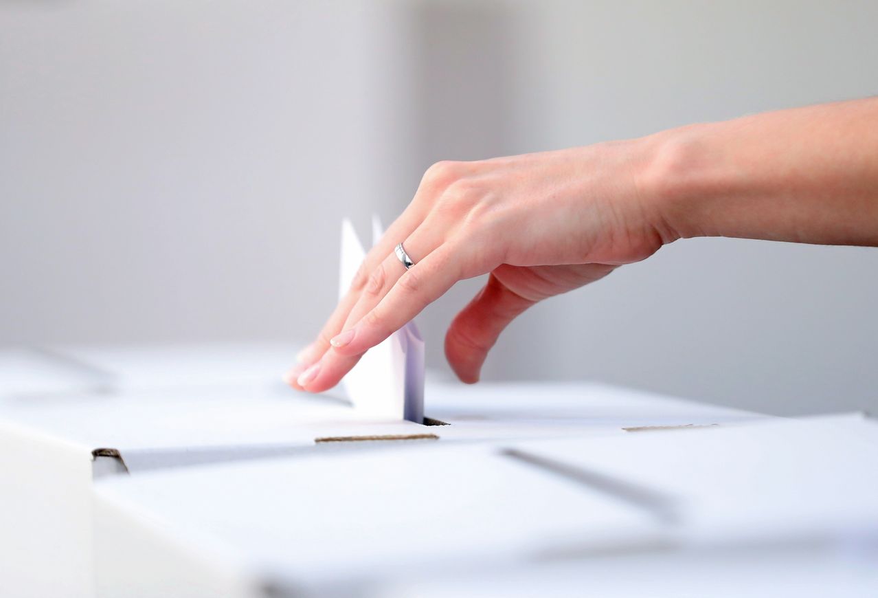 A close-up of a hand placing a folded paper into a white ballot box with a slit.