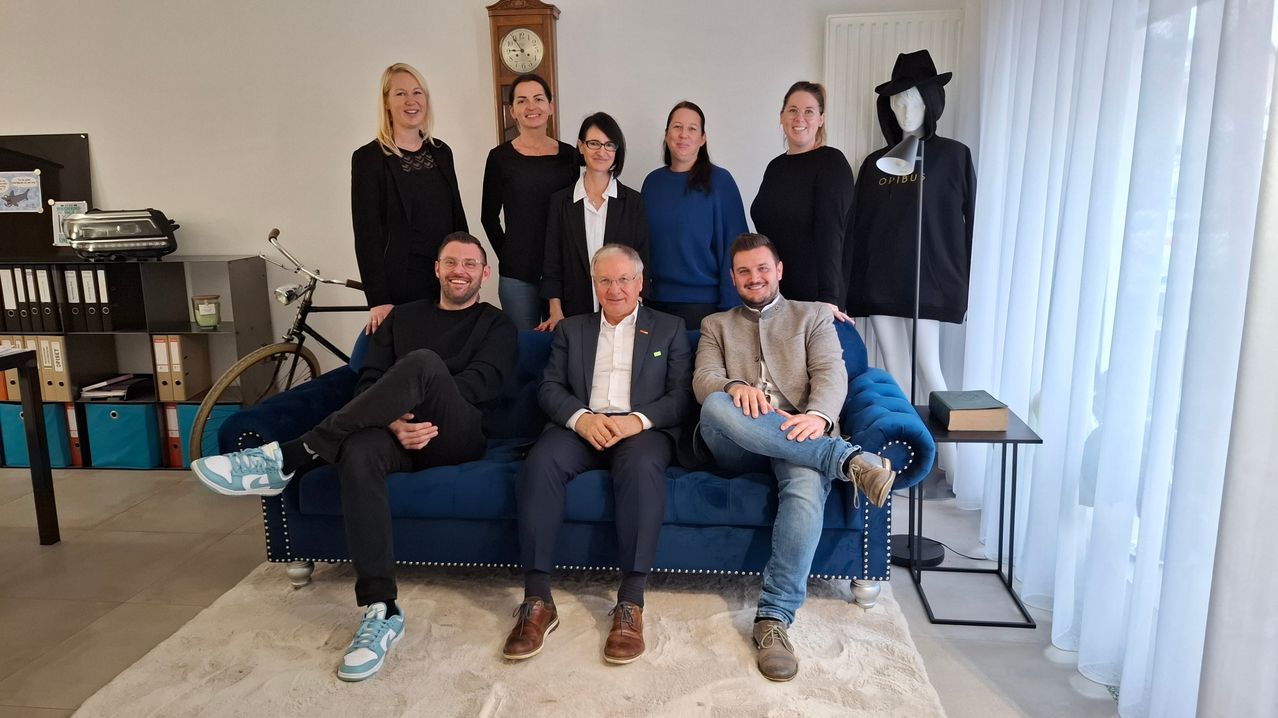 A group of people pose for a picture on a blue couch, with a clock and mannequin in the background.