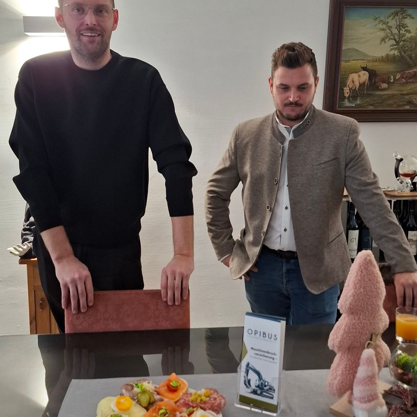 Two men stand in a dining room, smiling, with a table displaying a spread of food, a sign for OPIBUS, and decorative items. Behind them, a framed picture hangs on the wall.