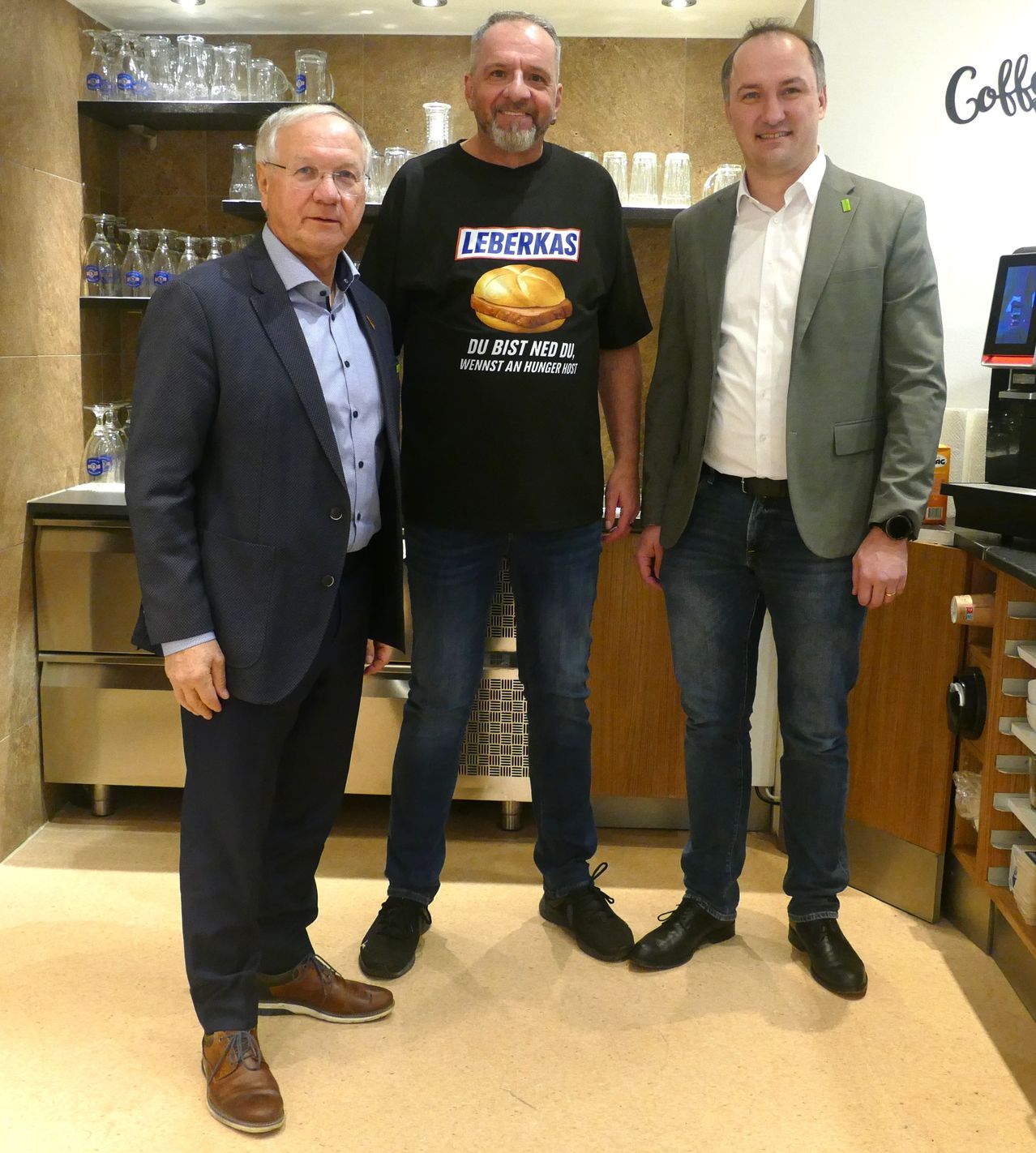Three men stand in a room with a coffee machine and shelves. One wears a black shirt with a hamburger design, and the other two wear formal suits.