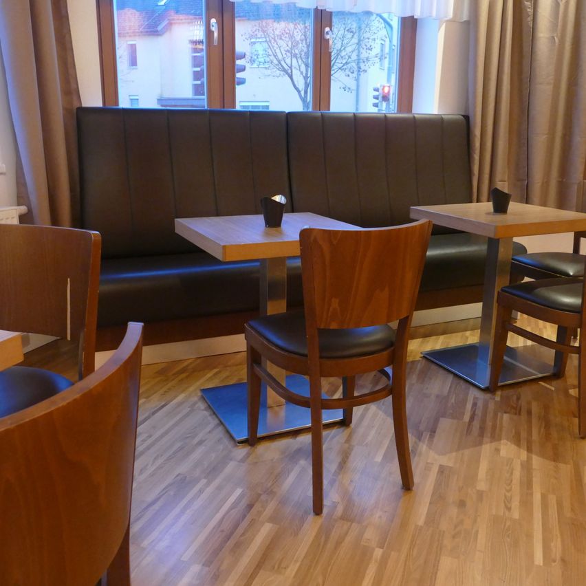 An indoor cafe features wooden tables and chairs. A brown leather bench seat is against the wall, and two tables have cups on them. Outside, buildings are visible through the glass window.