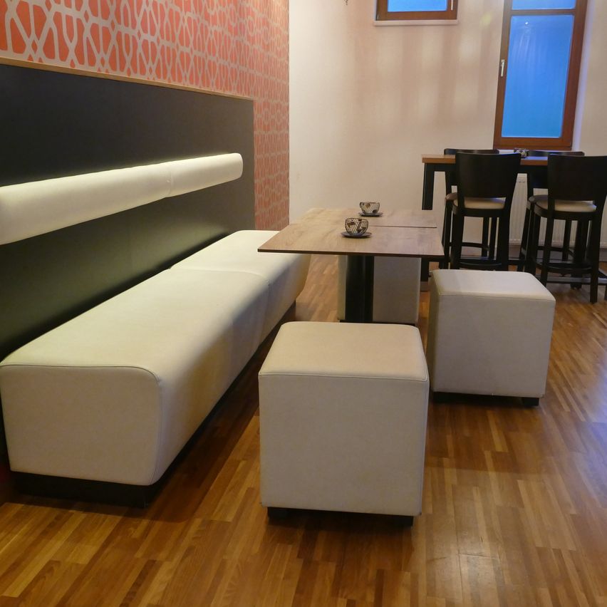 A dining area with a white bench, two stools, and a small wooden table. The room has wooden floors, orange and white walls, and a window.