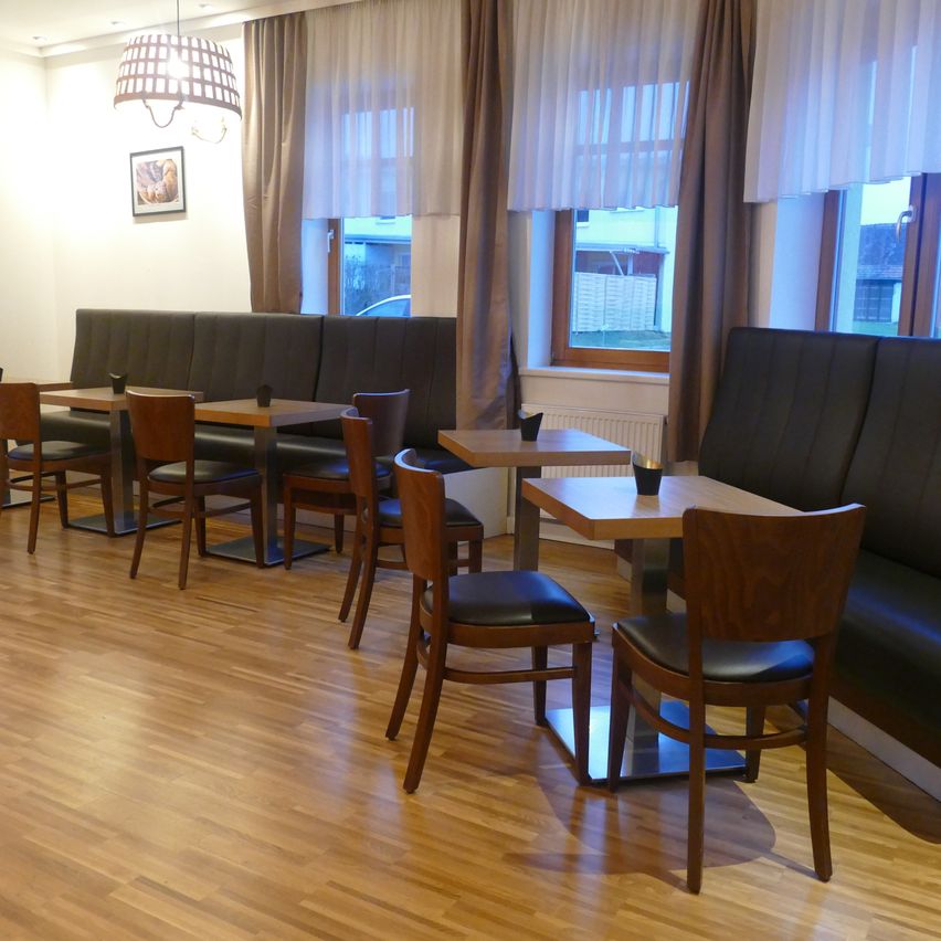 A restaurant interior with wooden floors, featuring cushioned seating around tables, draped windows, and a framed picture on the wall.